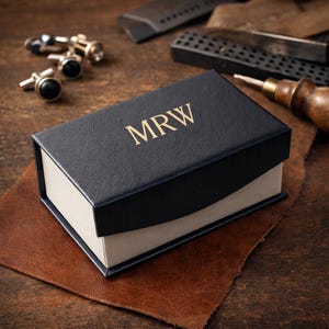 Puede incluir: Una caja de regalo azul marino con las iniciales "MRW" en letras doradas. La caja está sobre una superficie de cuero marrón, con varios pares de gemelos y herramientas en el fondo. El interior de la caja es blanco.