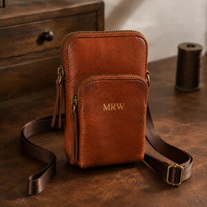 May include: A vertical brown leather crossbody bag with a zippered main compartment and a smaller front pocket. The front pocket features the embossed letters MRW. The bag includes a thin, adjustable dark brown fabric strap and antique brass hardware.