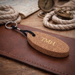 Op de afbeelding: Een ovale kurken sleutelhanger met de gouden initialen "TMH". Deze is bevestigd aan een zwart koord en een zilveren ring. De sleutelhanger ligt op een bruin leren oppervlak, met nautische elementen op de achtergrond.