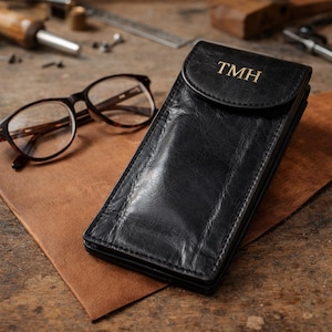 以下が含まれることがあります： 金色のイニシャル「TMH」が入った黒い革製メガネケース。べっ甲のメガネが、ケースの隣にある茶色の革パッドの上に置かれています。ケースはフラップで閉じられています。