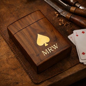 Peut inclure: Une boîte rectangulaire en bois brun foncé avec un couvercle à charnière. La surface supérieure présente un symbole de pique doré centré au-dessus des lettres MRW, également en or. La boîte a une finition lisse et un grain de bois visible.