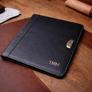 Peut inclure: Porte-documents en cuir noir avec des coutures dorées et un fermoir doré. Les initiales "TMH" sont estampées en or dans le coin inférieur droit. Le porte-documents est posé sur une surface en cuir marron.