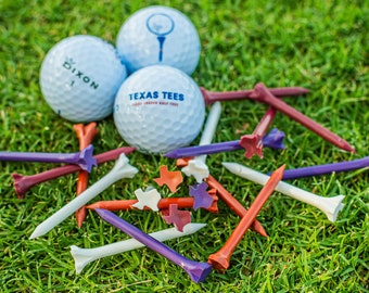 May include: Three white golf balls, one with the text "Dixon 1" and another with the text "Texas Tees" on a green grassy surface. There are multiple colorful golf tees scattered around the golf balls, including red, white, and purple tees with a Texas star design.