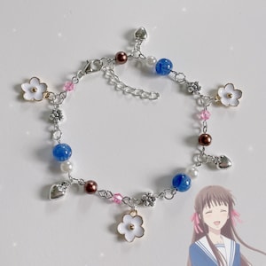May include: A silver chain bracelet with white flowers, blue and brown beads, and silver hearts. The bracelet is on a white background.