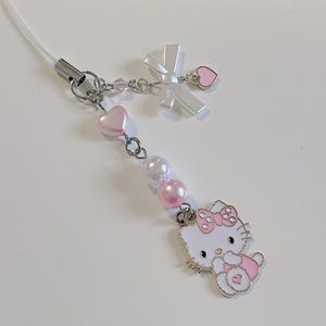 May include: A white Hello Kitty charm with pink accents and a white bow. The charm is attached to a silver chain with pink and white beads.