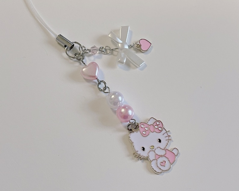 May include: A white Hello Kitty charm with pink accents and a white bow. The charm is attached to a silver chain with pink and white beads.