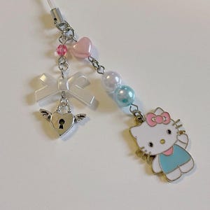 May include: A white and blue Hello Kitty charm with a pink bow, a silver heart with a keyhole, a white bow, and pink and blue beads. The charm is attached to a clear string.