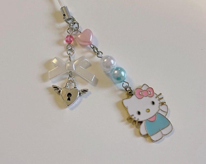 May include: A white and blue Hello Kitty charm with a pink bow, a silver heart with a keyhole, a white bow, and pink and blue beads. The charm is attached to a clear string.
