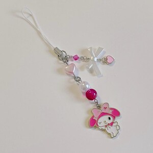 May include: A pink and white phone charm featuring a cartoon character with a pink bow and a white dress. The charm is attached to a white string with pink and white beads.