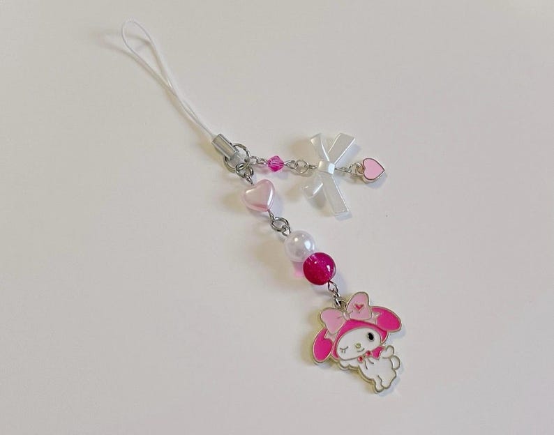 May include: A pink and white phone charm featuring a cartoon character with a pink bow and a white dress. The charm is attached to a white string with pink and white beads.