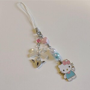May include: A white phone charm with a pink and blue Hello Kitty character, a white bow, a silver heart-shaped lock with wings, and pink and blue beads.