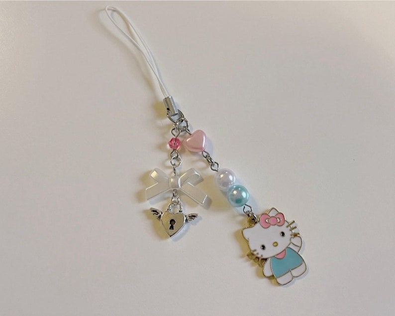 May include: A white phone charm with a pink and blue Hello Kitty character, a white bow, a silver heart-shaped lock with wings, and pink and blue beads.