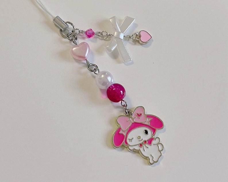 May include: A pink and white charm with a cartoon character wearing a pink hat and a pink bow. The charm is attached to a silver chain with pink and white beads and hearts.