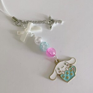 May include: A white and blue charm with a cartoon dog holding a cup, attached to a white string with a silver key charm, a white bow, and pastel blue and pink beads.