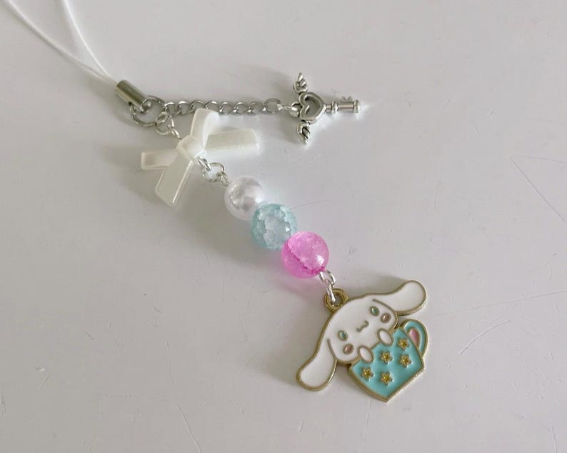 May include: A white and blue charm with a cartoon dog holding a cup, attached to a white string with a silver key charm, a white bow, and pastel blue and pink beads.