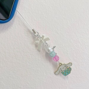 May include: A white phone charm with a white bow, pearl beads, and a blue and pink charm of a cartoon dog holding a cup.