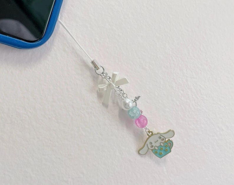 May include: A white phone charm with a white bow, pearl beads, and a blue and pink charm of a cartoon dog holding a cup.