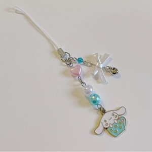 May include: A white phone charm with a cartoon dog holding a blue cup with stars. The charm has a white bow, pink and blue beads, and a silver bell.