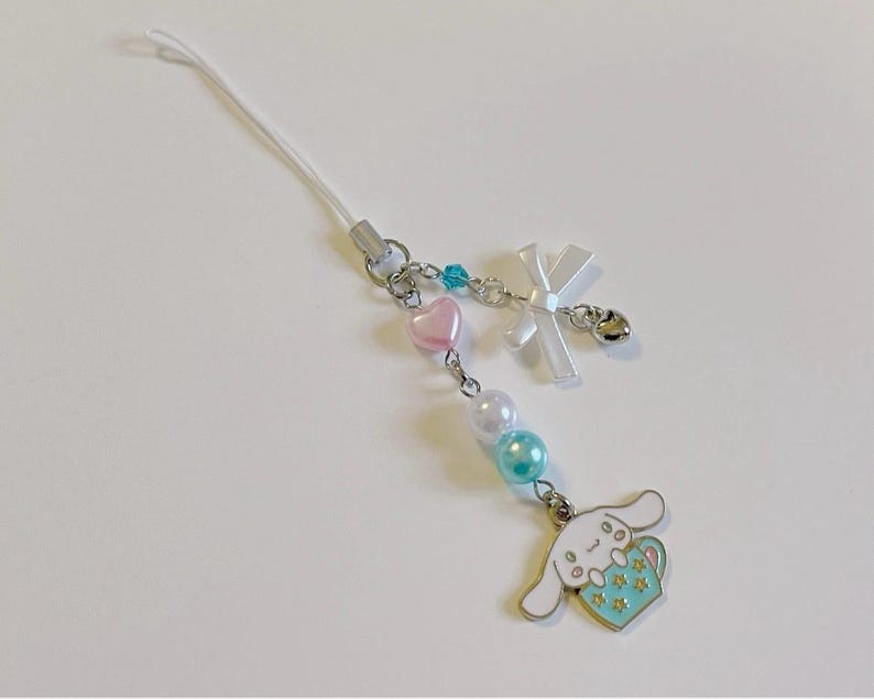 May include: A white phone charm with a cartoon dog holding a blue cup with stars. The charm has a white bow, pink and blue beads, and a silver bell.