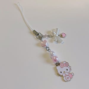 May include: A white and pink Hello Kitty phone charm with a white ribbon, pink hearts, and pearl beads.