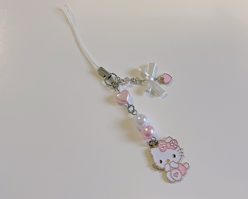 May include: A white and pink Hello Kitty phone charm with a white ribbon, pink hearts, and pearl beads.