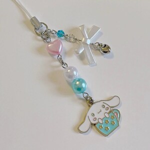 May include: A white phone charm with a pink heart, white pearls, and a turquoise blue charm. The charm features a cartoon dog holding a turquoise blue cup with gold stars.