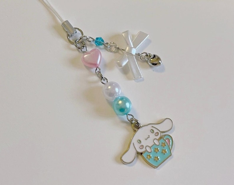 May include: A white phone charm with a pink heart, white pearls, and a turquoise blue charm. The charm features a cartoon dog holding a turquoise blue cup with gold stars.