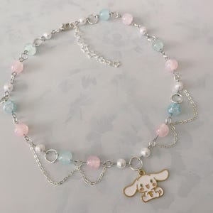 May include: A delicate necklace with pink, blue, and white beads, a silver chain, and a charm of a white cartoon character with gold accents. The necklace has a star-shaped clasp.