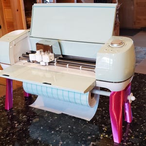 May include: A light blue and white crafting machine with a roll of white paper and pink legs. The machine has a lid that is open and a silver cutting blade.