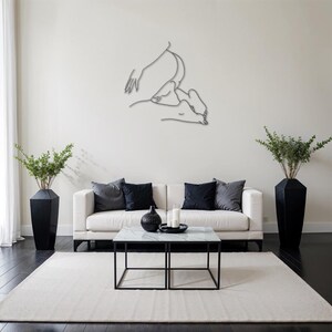 May include: A white couch with black throw pillows sits in a living room with a white rug. A black metal coffee table with a white marble top sits in front of the couch. A black vase with green leafy plants sits on the floor to the right of the couch. A black and white line drawing of two figures is hanging on the wall behind the couch.