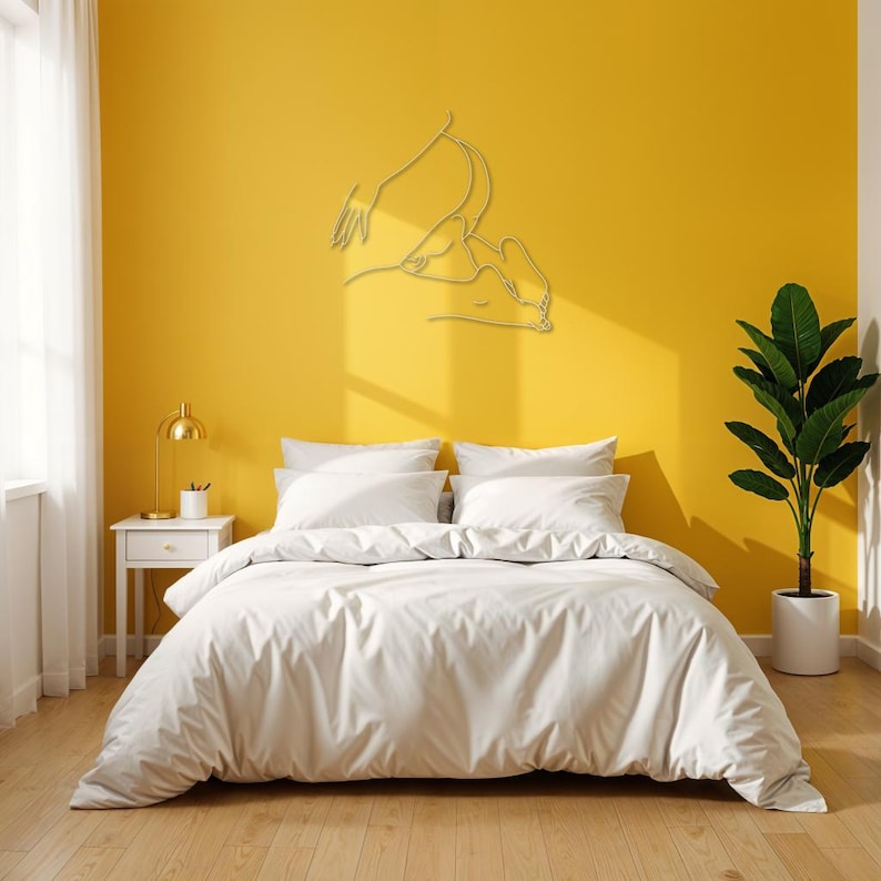 May include: A white bed with a white duvet cover and two white pillows sits in front of a yellow wall. A gold metal line art sculpture of a couple is on the wall above the bed. A small gold lamp sits on a white nightstand next to the bed. A potted green plant is on the right side of the image.