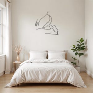 May include: A white bed with a white duvet cover and two white pillows. A metal line art sculpture of a couple embracing is hanging on the wall above the bed.
