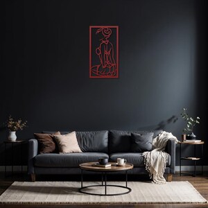 Puede incluir: Una escultura de pared de metal rojo que representa a una mujer sentada. La escultura est&aacute; montada en una pared gris oscuro sobre un sof&aacute; gris con almohadas y una manta.