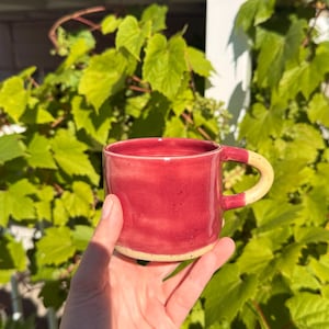 Peut inclure: Une tasse en céramique faite à la main avec un vernis magenta vif sur le corps principal et une anse contrastée de couleur crème mouchetée. La base de la tasse présente également cette finition crème mouchetée, créant un aspect bicolore.