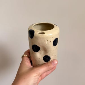 Peut inclure: Un récipient cylindrique en céramique avec une base couleur crème, des taches brun foncé et de grands pois noirs irréguliers. La surface présente une légère indentation et une petite ouverture en bec verseur triangulaire.