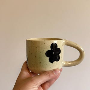 May include: A cream-colored ceramic mug with a speckled texture and a hand-painted black flower design on the side. The mug features a rounded handle and a cylindrical body, showcasing a simple, functional design with a smooth, glazed finish.