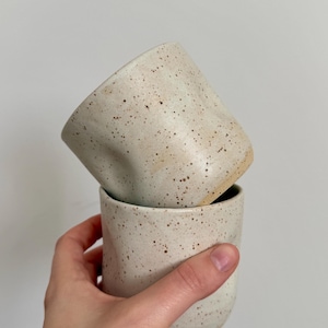 Könnte beinhalten: Zwei gesprenkelte Keramikbecher, einer auf dem anderen gestapelt. Die Becher haben eine neutrale Farbe mit dunkelbraunen Sprenkeln. Der obere Becher ist leicht angewinkelt und gibt den Blick ins Innere frei. Die Becher werden von einer Hand gehalten.