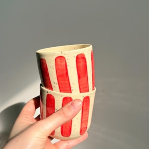 Peut inclure: Deux tasses en céramique empilées avec une base de couleur crème présentant des détails mouchetés et des rayures verticales peintes en rouge. Les tasses ont une forme arrondie lisse sans anse, avec un aspect peint à la main.