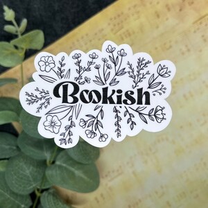 May include: A white sticker with the word "Bookish" in a bold, black font, surrounded by delicate black floral illustrations. The sticker has a scalloped edge and is set against a textured, light-colored background with green foliage.