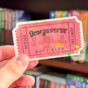 May include: A pink holographic sticker with the text "Omegaverse Book Club" and a rope knot with the Greek letters alpha and beta. The sticker has the words "ADMIT ONE" on the right side.