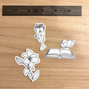 Puede incluir: Tres pegatinas de contorno blanco sobre una superficie de madera. Una pegatina representa a una persona leyendo con flores, otra una flor de lirio y la última un libro abierto con un corazón. Una regla está en la parte superior.