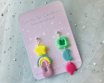 May include: A pair of handmade dangle earrings featuring a rainbow, star, moon, shamrock, and heart charm. The earrings are on a white card with the text "Castles & Cauldrons Handcrafted".