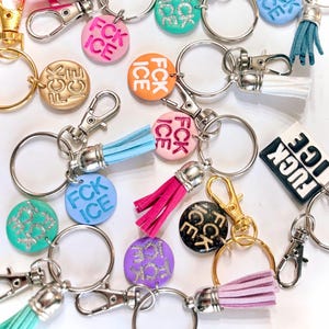May include: A collection of keychains with round charms and tassel accents. The charms feature the text "FCK ICE" in various colors, including pink, orange, and blue. Keychains have silver and gold hardware.