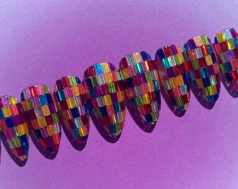 May include: A set of nine artificial nails, each with a colorful, mosaic-like design. The nails are arranged in a row against a purple background. The mosaic pattern is made up of small, rectangular pieces in various colors.