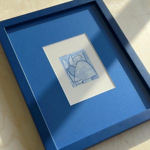 May include: A framed artwork featuring a blue abstract design on white paper. The artwork is set within a blue mat and a matching blue frame. The piece is displayed on a light wood surface.