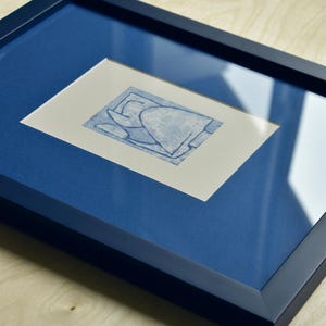May include: A framed abstract art print. The artwork features a blue design on a white background, set within a blue mat and a dark blue frame. The frame is rectangular and has a glossy finish. The artwork is displayed on a light wood surface.