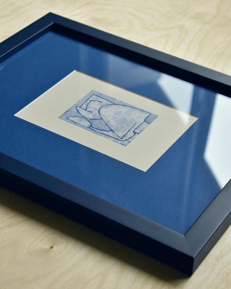 May include: A framed abstract art print. The artwork features a blue design on a white background, set within a blue mat and a dark blue frame. The frame is rectangular and has a glossy finish. The artwork is displayed on a light wood surface.