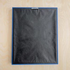 May include: Back of a framed artwork with a blue border. The frame is rectangular, with a black backing and a metal sawtooth hanger at the top for wall mounting. The frame is set against a light wood surface.