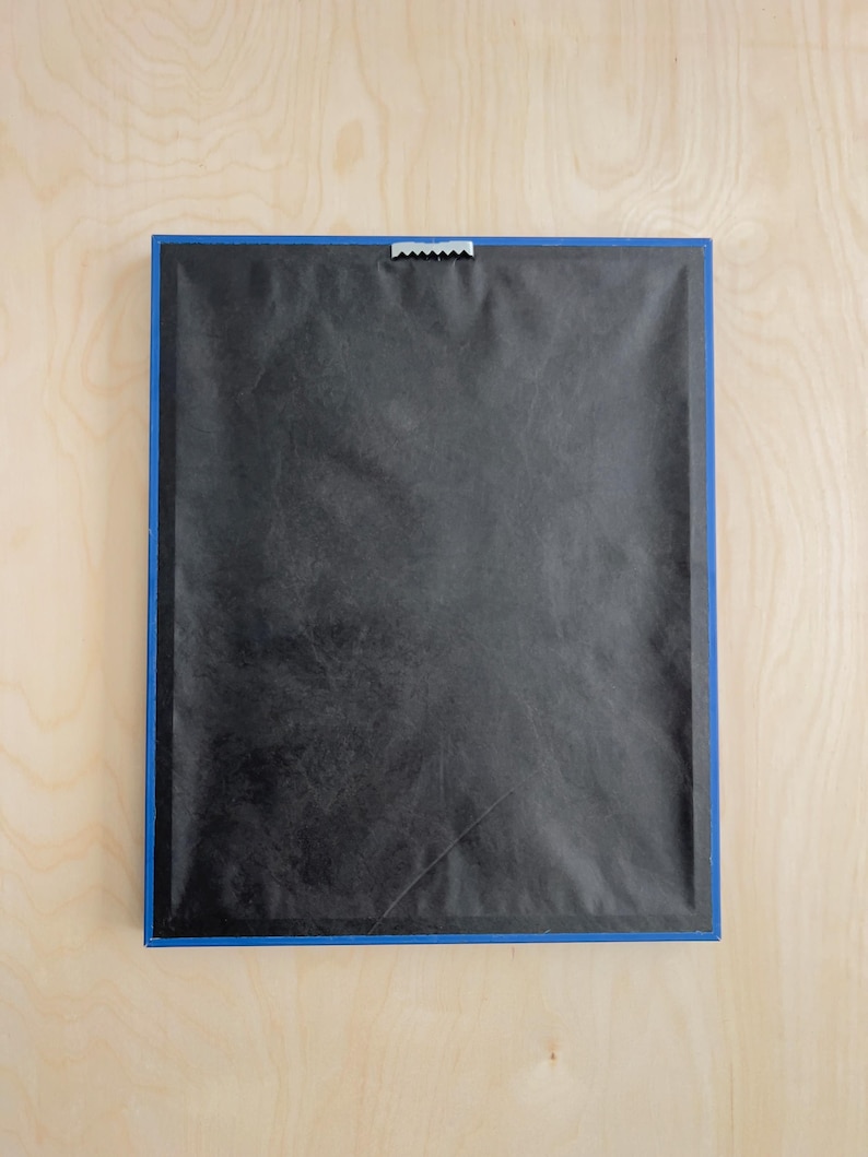 May include: Back of a framed artwork with a blue border. The frame is rectangular, with a black backing and a metal sawtooth hanger at the top for wall mounting. The frame is set against a light wood surface.