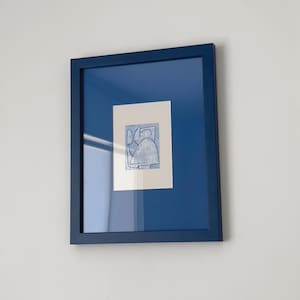 May include: A framed abstract art print. The artwork features a light blue design on a white background, set within a deep blue mat and frame. The piece is mounted on a white wall, creating a clean and modern aesthetic.